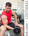 Man in his 20s performing seated dumbbell curl on gym bench wearing red sleeveless shirt 135716422