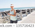 Mature woman walking by concrete rail on paved path, wearing white visor and leggings, rocky shore 135716423