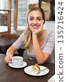 Woman in mid-20s sitting at cafe table wearing light gray top, holding cup, plate with cake 135716424