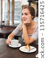 Woman sitting at coffee shop, resting chin on left hand, touching white cup on wooden table 135716425