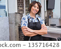 Mature female stylist standing arms crossed inside salon wearing black apron with scissors comb 135716428