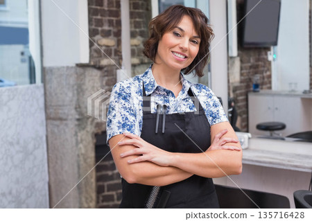 Mature female stylist standing arms crossed inside salon wearing black apron with scissors comb 135716428