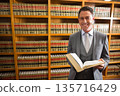 Man in suit standing by wooden bookshelves in law library holding open legal volume, copy space 135716429