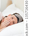 Woman lying on bed with white pillow and bedside lamp, wearing gray sleep mask, covering mouth 135716430