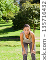 Adult female jogging, pausing and leaning with hands on knees in sunlit park, wearing gray leggings 135716432