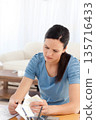 Woman sitting at wooden table wearing blue top in living room, sorting bills, using calculator 135716433