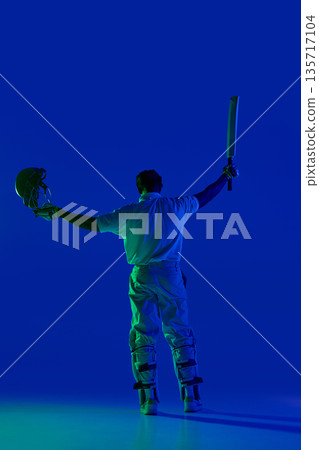 Cricket batsman standing with raised equipment under blue studio light 135717104