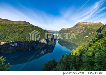 Aerial panoramic view of Geirangerfjord in summer Norway. Scenic panoramic landscape of Geirangerfjord with steep green mountains and calm blue water under clear summer sky in Norway. 135719305