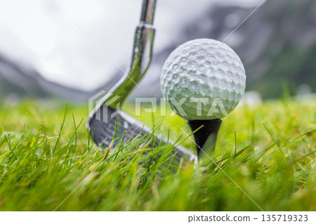 Golf club and ball on wet green field with mountains in background. 135719323