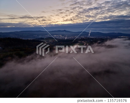 aerial shot portraying sunrise through clouds over misty valley with glowing city and industrial steam 135719376