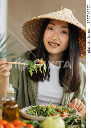 A young Asian woman wearing a conical hat enjoys a healthy salad with chopsticks, surrounded by fresh ingredients and olive oil 135719771