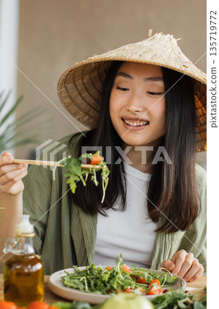 A young Asian woman wearing a conical hat enjoys a fresh salad with chopsticks, surrounded by healthy ingredients and olive oil A young Asian woman wearing a conical hat enjoys a fresh salad with chopsticks, surrounded by healthy ingredients and olive oil 135719772