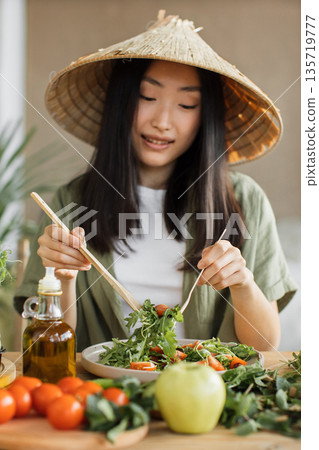 A young Asian woman wearing a conical hat prepares a fresh salad with tomatoes and greens, showcasing healthy eating habits 135719777