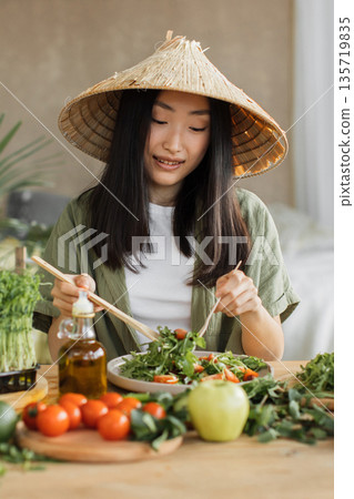 A young Asian woman wearing a conical hat enjoys a fresh salad with chopsticks, surrounded by healthy ingredients 135719835
