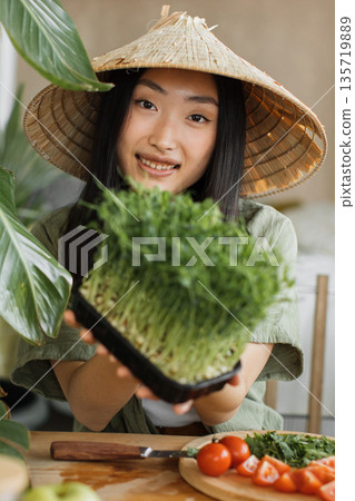 A young woman wearing a conical hat proudly displays a tray of freshly grown microgreens, surrounded by fresh vegetables on a wooden table A young woman wearing a conical hat proudly displays a tray of freshly grown microgreens, surrounded by fresh vegetables on a wooden table 135719889