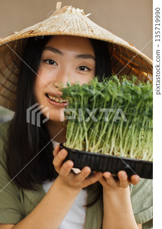 A young woman wearing a conical hat holds a tray of fresh microgreens, smiling warmly at the camera 135719990