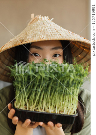 A young woman wearing a conical hat holds a tray of fresh microgreens in front of her face, showcasing healthy eating and sustainable agriculture 135719991