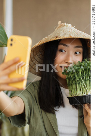 A young Asian woman wearing a conical hat takes a selfie with a tray of fresh microgreens in her hand 135719992