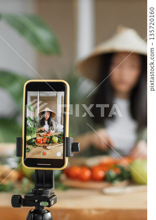 A young Asian woman wearing a conical hat films herself preparing fresh vegetables for a cooking tutorial on her smartphone 135720160