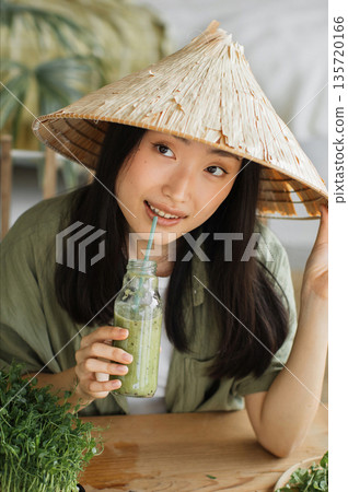 A young Asian woman wearing a conical hat enjoys a healthy green smoothie from a glass bottle with a straw, surrounded by fresh greens A young Asian woman wearing a conical hat enjoys a healthy green smoothie from a glass bottle with a straw, surrounded by fresh greens 135720166