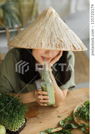 A young woman wearing a conical hat sips a healthy green smoothie from a glass bottle with a straw, surrounded by fresh ingredients on a wooden table A young woman wearing a conical hat sips a healthy green smoothie from a glass bottle with a straw, surrounded by fresh ingredients on a wooden table 135720174