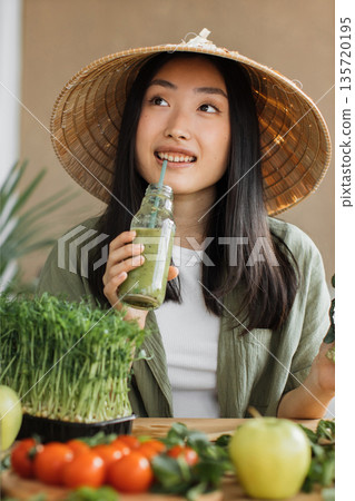A young Asian woman wearing a conical hat drinks a healthy green smoothie surrounded by fresh vegetables and microgreens A young Asian woman wearing a conical hat drinks a healthy green smoothie surrounded by fresh vegetables and microgreens 135720195