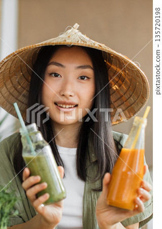 A young Asian woman wearing a traditional conical hat holds two bottles of colorful juice, one green and one orange, ready for a healthy refreshment A young Asian woman wearing a traditional conical hat holds two bottles of colorful juice, one green and one orange, ready for a healthy refreshment 135720198