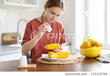 Young Girl Salting Boiling Corn Cobs For Eating In The Kitchen Young Girl Salting Boiling Corn Cobs For Eating In The Kitchen 135720769