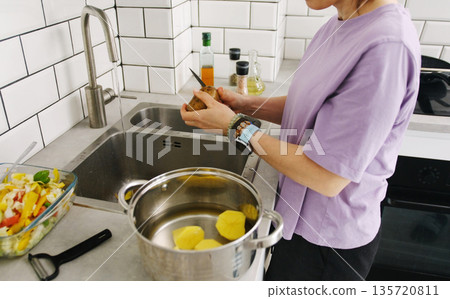 Woman Peeling A Potato In The Kitchen And Cutting It Into A Pot Woman Peeling A Potato In The Kitchen And Cutting It Into A Pot 135720811