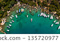 Aerial view of small boats and yachts anchored in a turquoise bay with clear water, wooden piers, rocky shoreline, and lush green trees. Summer seaside marina from drone. 135720977