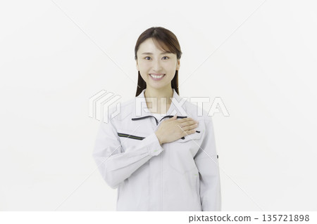 A young Japanese woman in work clothes holding her hand on her chest against a white background A young Japanese woman in work clothes holding her hand on her chest against a white background 135721898