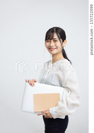 A young woman smiling while looking at the camera and holding a laptop and notebook in front of a white background A young woman smiling while looking at the camera and holding a laptop and notebook in front of a white background 135722099