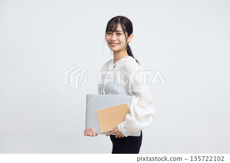A young woman smiling while looking at the camera and holding a laptop and notebook in front of a white background A young woman smiling while looking at the camera and holding a laptop and notebook in front of a white background 135722102