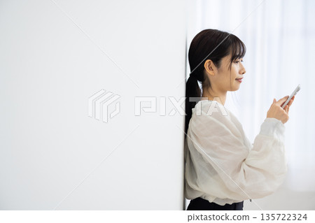 Profile of a young woman leaning against a white wall and operating a smartphone Profile of a young woman leaning against a white wall and operating a smartphone 135722324