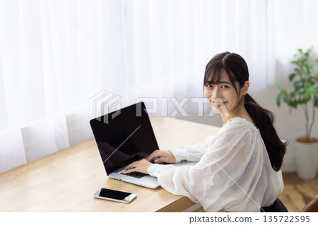 A young woman smiling at the camera while using a computer at a desk by the window 135722595