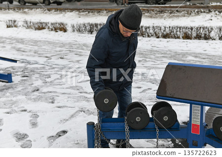 Man exercises outdoors on gym equipment in winter in a city park Man exercises outdoors on gym equipment in winter in a city park 135722623