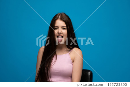 Playful young woman making funny face with tongue out against blue background, humor and carefree emotion concept 135723890