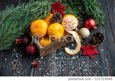 Orange mandarins in wooden box with fir apple, red balls, snowflakes on green fir branch background. Christmas card. Happy new year and merry christmas. High quality photo 135724940