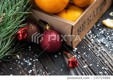 Orange mandarins in wooden box with fir apple, red balls, snowflakes on green fir branch background. Christmas card. Happy new year and merry christmas. High quality photo 135724966
