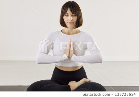 Serene woman practicing yoga meditation in lotus with prayer hands indoors 135725654