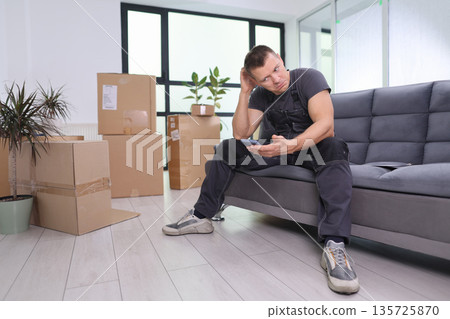 Moving day stress as a man sits on a couch with boxes, looking at his phone in a bright, modern living room during the afternoon Moving day stress as a man sits on a couch with boxes, looking at his phone in a bright, modern living room during the afternoon 135725870