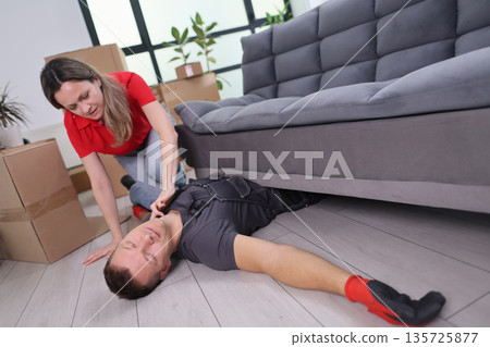 Efforts to rescue a fallen mover trapped beneath a couch in a bright, modern living room filled with moving boxes in the afternoon 135725877
