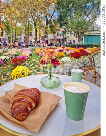 Charming outdoors cafe scene during autumn. A white marble table holds a delicious croissant with two coffee cups and a vase of colorful seasonal flowers. People enjoying the warm, inviting atmosphere Charming outdoors cafe scene during autumn. A white marble table holds a delicious croissant with two coffee cups and a vase of colorful seasonal flowers. People enjoying the warm, inviting atmosphere 135726056