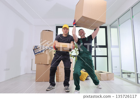 Hardworking movers lift boxes with smiles in a spacious, bright room filled with packing materials and cardboard cartons Hardworking movers lift boxes with smiles in a spacious, bright room filled with packing materials and cardboard cartons 135726117