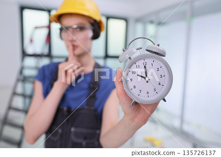 Focused builder displays clock while signaling silence in a busy construction zone during work hours Focused builder displays clock while signaling silence in a busy construction zone during work hours 135726137