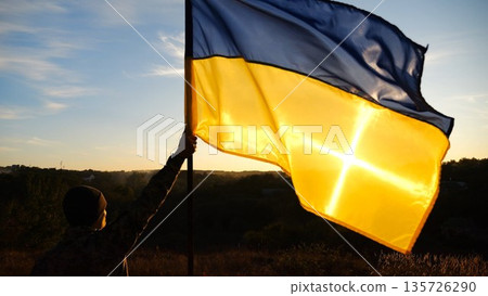 Young soldier of ukrainian army lifted blue-yellow banner in honor of the victory against russian aggression at sunset. Male military in uniform raised a waving flag of Ukraine at countryside Young soldier of ukrainian army lifted blue-yellow banner in honor of the victory against russian aggression at sunset. Male military in uniform raised a waving flag of Ukraine at countryside 135726290