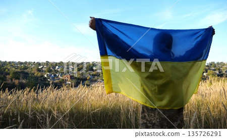 Military man in uniform stands with raised over head flag of Ukraine at countryside. Male ukrainian army soldier with lifted national banner in honor of victory against russian aggression. End of war Military man in uniform stands with raised over head flag of Ukraine at countryside. Male ukrainian army soldier with lifted national banner in honor of victory against russian aggression. End of war 135726291