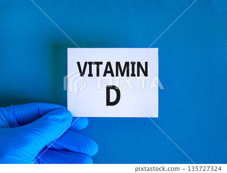 Vitamin D symbol. Concept words Vitamin D on beautiful white card. Doctor's hand in blue medical glove. Beautiful blue background. Medical healthy lifestyle Vitamin D concept. Vitamin D symbol. Concept words Vitamin D on beautiful white card. Doctor's hand in blue medical glove. Beautiful blue background. Medical healthy lifestyle Vitamin D concept. 135727324