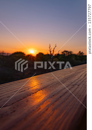 Golden sunset light reflecting on wooden railing. Serene nature scene with sun low over horizon. Warm evening glow, peaceful outdoor view. 135727797
