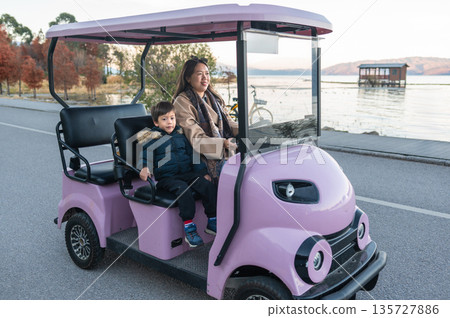 Mother And Child Riding A Pink Electric Vehicle By The Lake Shore. Mother And Child Riding A Pink Electric Vehicle By The Lake Shore. 135727886
